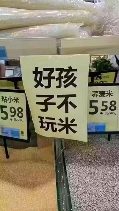 这是超市粮油散装区的实拍场景，中间张贴着一张手写趣味标语「好孩子不玩米」，左侧是粘小米的价格牌，标价5.98元/500g，下方对应着黄色的粘小米；右侧是荞麦米的价格牌，标价5.58元/500g，下方对应着灰色的荞麦米。
标语采用直白的口语化劝诫语气，以类似教育小朋友的方式提醒顾客不要随意触碰、拨弄散装粮食，风格幽默接地气，让人忍俊不禁。