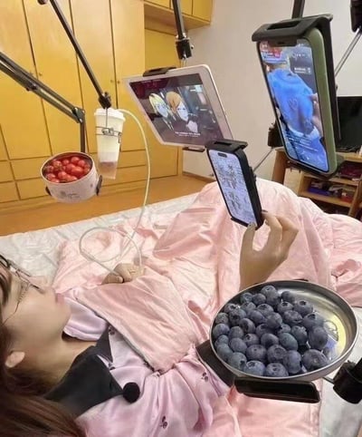 这张图片展示了一位女生全副武装地躺在床上，身旁和床头装满了支架，分别用来固定零食、水果、饮料、手机和平板。她一边吃着蓝莓，吸着饮料，同时用不同的设备看直播、动漫等内容，体现了极致的懒宅生活方式。画面非常夸张，带有明显的搞笑和自嘲色彩，暗示着现代人宅家生活的舒适与效率最大化，并未涉及低俗或明显暗示。

值得注意的是图中色彩丰富，对自动化懒人设备进行了一种幽默的极致演绎。此类场景常在年轻人社交网络广泛传播，具有自调侃双关意味，甚至有“现代版葛优躺”的味道。
