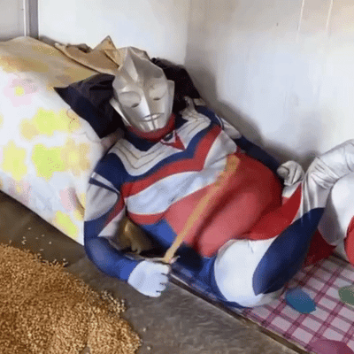 一位穿着奥特曼服装的人，挺着夸张的大肚子，悠闲地躺在地上。他手持一根木棍，周围散落着谷物，似乎在享受悠闲的时光。这个场景通过奥特曼这一经典英雄形象与慵懒姿态的反差，创造出幽默搞笑的效果，反映了现代生活中人们对放松和