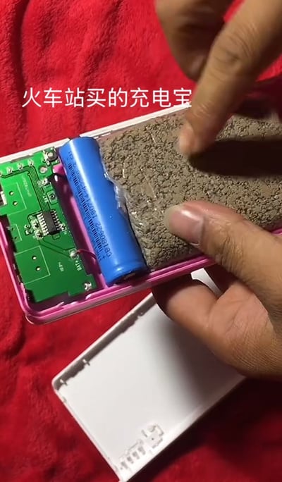 图片展示了一个被拆开的粉色充电宝，内部结构暴露在外。左侧可见绿色的电路板和一节蓝色的圆柱形电池，而电池右侧的大部分空间被灰色的沙子或混凝土填充。一只手的手指正指向填充的沙子部分，似乎在强调这一发现。图片上方有白色文字标注“火车站买的充电宝”，揭示了该充电宝的购买地点。整体场景直观地展示了假冒伪劣充电宝的内部构造，通过填充沙子来增加重量以冒充正规产品。