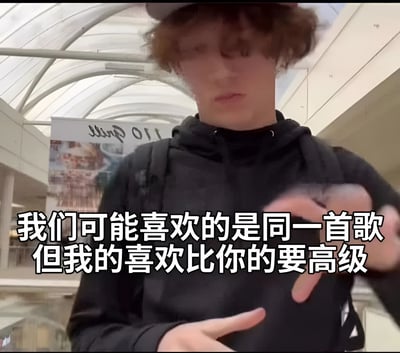 图片中是一名卷发男子，他戴着帽子，穿着黑色连帽衫，背着黑色背包，站在一个有玻璃穹顶的商场走廊里。男子面无表情，双手在胸前比出特定手势。图片下方有一行白色文字："我们可能喜欢的是同一首歌但我的喜欢比你的要高级"，表达了一种音乐品味上的优越感。