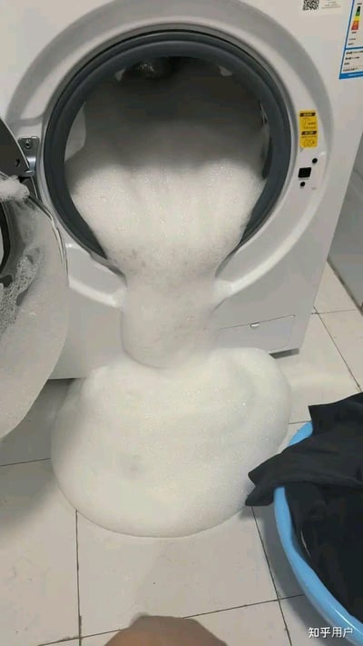 这是一张真实生活场景实拍图，画面中的滚筒洗衣机因洗涤剂投放过量，大量白色泡沫从舱门涌出，堆积在白色瓷砖地面上，旁边还放着一个装有深色衣物的蓝色水盆，呈现出洗衣时的翻车小意外，让人感到无奈又哭笑不得。