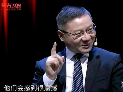这是一张东方卫视节目的视频截图，画面中是学者张维为，他身着深色西装、白衬衫搭配蓝白条纹领带，佩戴眼镜，耳挂式麦克风，面带自信的笑容，右手食指向上指，正在发表观点。图片底部配有白色字幕“他们会感到很震撼”，该画面被截取后成为网络表情包，常被用于社交平台，用来表达对某件事的惊叹、强调情绪，或是作为调侃的素材。