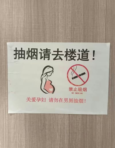 这是一张实拍的提示标识照片，标识张贴在带有木纹纹理的浅色墙面上。标识顶部用黑色粗体大字写着“抽烟请去楼道！”，中部左侧是一个长发孕妇的剪影图标，孕妇孕肚处还有一个白色爱心图案，中部右侧是标准的红圈斜杠禁止吸烟标识，标识下方标注了红色文字“禁止吸烟”以及英文“NO SMOKING”，标识最底部用红色字体写着提示语“关爱孕妇 请勿在男厕抽烟！”，这条标语因为逻辑反常显得滑稽，孕妇极少出现在男厕，该标语的设定不符合日常场景，让人疑惑。