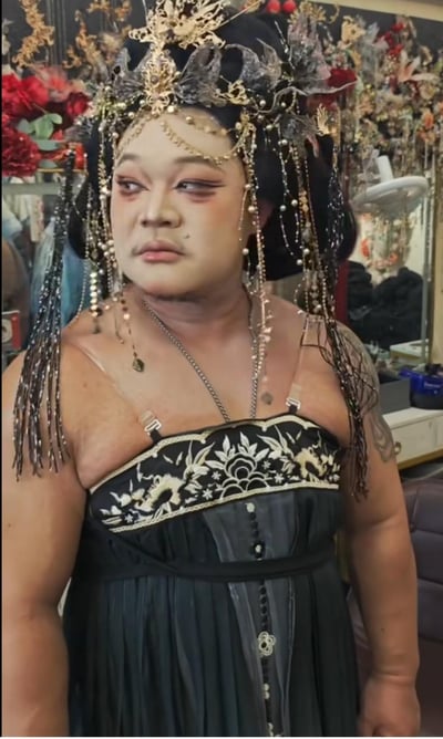 图中是一位身材壮硕的男性，化着精致的古风女性妆容，脸部的小胡子与柔美的妆容形成强烈视觉反差。他头戴华丽繁复的黑金系古风头饰，垂挂着大量珠链与流苏，身穿带有精致白色刺绣的黑色低胸古风吊带裙，露出的肩膀和手臂带有纹身，身处摆放着镜子、装饰花的化妆间场景中。整体造型将壮汉的硬朗与古风女性造型的柔美结合，充满反差感，十分搞笑吸睛。图中无谐音梗、双关或暗示内容。