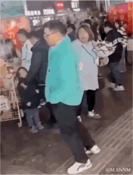 这是一段在热闹的室内公共场合（疑似集市或商场）拍摄的动态画面，一位穿着亮绿色外套的大叔迈着夸张魔性的步伐行走，周围的路人纷纷将目光投向他，画面充满搞笑氛围，大叔的走路姿势成为视觉焦点，显得十分滑稽有趣。