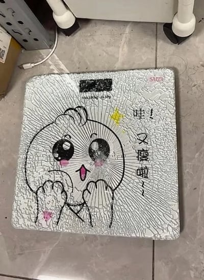 这是一张实拍的静态照片，拍摄的是放置在浅灰色瓷砖地面上的正方形玻璃体重秤，秤的玻璃面板已经完全碎裂，布满了密集的蛛网状裂纹。秤面原本印有可爱的Q版卡通圆脸图案，脸颊带有粉色腮红，睁着大眼睛做出双手捧脸的惊喜姿态，图案旁配有正向鼓励的文案“哇！又瘦啦~”，秤面上方还有电子称重显示区域，标注了最大称重180kg、精度50g的参数，右上角印有品牌标识。碎裂的秤面和原本温馨鼓励的减重文案形成强烈反差，暗含“体重太重把秤压碎”的调侃笑点，极具搞笑效果。