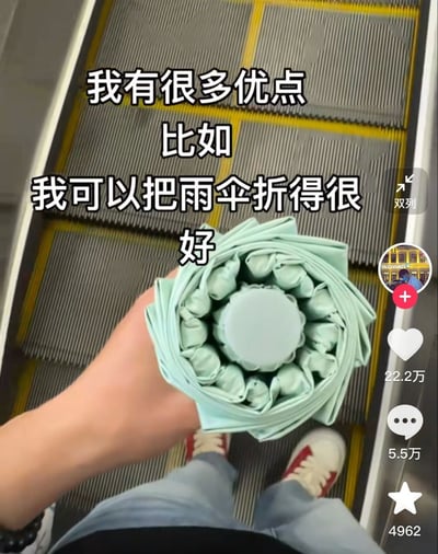 这是一张第一视角拍摄的短视频平台截图，拍摄者正站在带有黄色警示线的自动扶梯上，左手举着一把折叠得极为整齐的浅绿色雨伞进行展示，雨伞收折后的褶皱均匀规整，视觉上十分治愈。拍摄者身穿浅蓝色牛仔裤，脚蹬白红拼色运动鞋。画面上方叠加有带黑色描边的白色配文，内容为“我有很多优点 比如 我可以把雨伞折得很好”。画面右侧是抖音的功能按钮栏，显示该内容获22.2万点赞、5.5万条评论、4962次收藏，还带有发布者的头像和关注按钮。