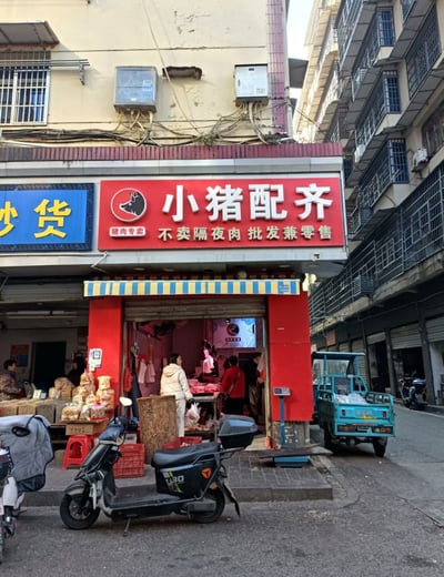 图片展示了老城区街道的一处猪肉专卖店场景。店铺招牌为红色底色，醒目地写着“小猪配齐 猪肉专卖”，下方小字标注“不卖隔夜肉 批发兼零售”，左侧相邻店铺有蓝色“炒货”招牌。店门口有红色门面和蓝黄条纹遮阳棚，店内有顾客正在挑选猪肉，店外停放着一辆蓝色三轮车，周围是带有铁栏杆和空调外机的老旧居民楼，整体呈现出生活化的社区市场氛围。

**谐音梗说明**：店铺名称“小猪配齐”谐音热门卡通形象“小猪佩奇（Peppa Pig）”，利用谐音吸引顾客注意，具有幽默效果。