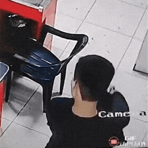 这是一段监控视角的动态GIF图，画面呈现室内场景，地面是浅色瓷砖，一旁设有红色柜台。一名扎马尾的人背对着镜头站立，其身旁的蓝色塑料椅子毫无征兆地向柜台方向快速滑动，看起来仿佛“自己动了起来”，营造出类似灵异事件的错觉，实际大概率是视觉错位或被隐藏的外力触发，画面充满了诡异又搞笑的反差感。