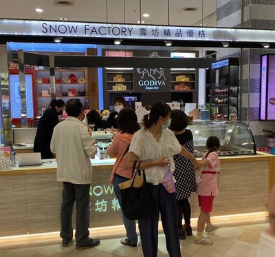 商场内一家名为“SNOW FACTORY 雪坊精品優格”的店铺前，有多位顾客正在排队购买商品。画面中可见不同年龄段的顾客，包括成年人和一名儿童，他们站在柜台前，部分人正在与店员交流。店铺柜台后方有货架，摆放着各类商品，背景中还能看到“GODIVA”的品牌标志，显示该店铺可能位于商场的美食区或品牌集中区域。顾客们大多佩戴着口罩，整体场景呈现出日常购物的热闹氛围。