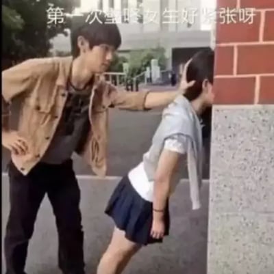 整张图为真人实拍素材配文的搞笑梗图，顶部配白色文字“第一次壁咚女生好紧张呀”，画面左侧是一名穿卡其色外套、黑色裤子的男性，他一只手叉腰，另一只手按在右侧人物的后脑勺，将对方按向墙面，做出壁咚的姿势；被按的人物穿着白短袖、深蓝色百褶裙，看似女生，但从身形和发型能看出实际是男性，刻意营造出与配文描述不符的反差感，笑点突出，用来调侃情感互动里的乌龙、尴尬或者出人意料的状况。
