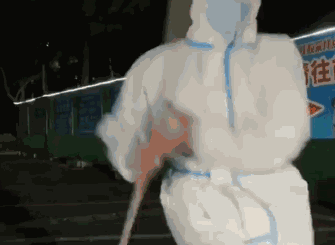 这是一段动态GIF，展示了夜晚户外街头，一位身着白色带蓝边防护服的防疫工作人员，手持类似扫帚的物品，正在欢快地跳舞，动作活泼充满喜感。背景能看到部分店铺招牌，整体氛围轻松诙谐，记录了防疫工作者在工作之余放松的瞬间。