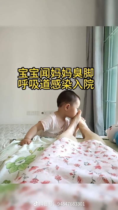 这是一张竖屏的室内床上场景实拍图，画面里一个小宝宝趴在床上，凑近闻一位成人的脚部，画面上方配有黄色加粗的警示文字“宝宝闻妈妈臭脚 呼吸道感染入院”，以此向家长传递警示信息：不注意卫生的亲子互动可能会导致宝宝患上呼吸道疾病，需重视育儿过程中的卫生问题。