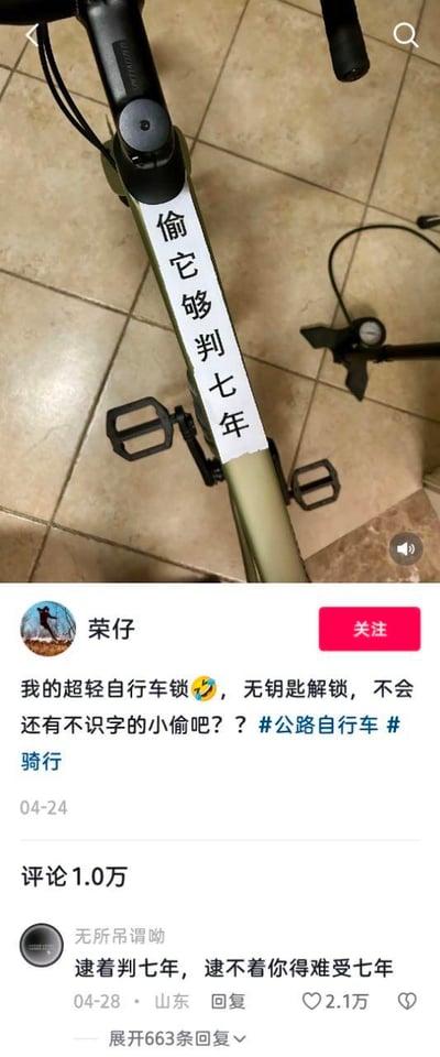 图片上半部分是自行车车架特写，车架上贴着一张竖排白色纸条，黑色字体写着“偷它够判七年”。下半部分是社交媒体帖子截图，发布者“荣仔”配文“我的超轻自行车锁😎，无钥匙解锁，不会还有不识字的小偷吧？？#公路自行车 #骑行”，发布时间04-24。帖子下方显示评论1.0万，其中高赞评论来自“无所谓嘞呦”：“逮着判七年，逮不着你得难受七年”，获赞2.1万，发布于04-28山东。整体通过幽默标语模拟“防盗锁”功能，调侃小偷识字问题，评论区形成趣味互动。