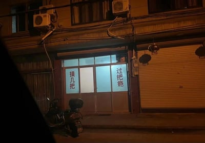 夜间拍摄的老旧街区店铺照片。画面中央是一家亮着灯的小店，玻璃门上贴着两组红色竖排标语，左侧为“摸几把”，右侧为“过把瘾”。店铺上方有杂乱的电线和空调外机，右侧是关闭的卷帘门，门前停放着一辆摩托车。整体氛围呈现出市井生活的烟火气，招牌文字组合略显诙谐，可能引发观者联想。
