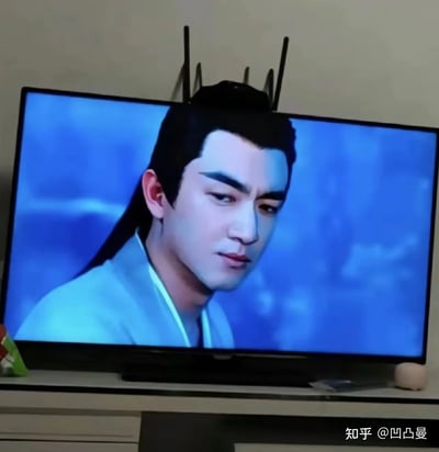 图片中展示了一台电视正在播放古装剧画面，屏幕上是一位身着浅蓝色古装、梳着长发的男性角色，背景为蓝色云雾。电视顶部摆放着几根黑色天线，从特定角度看过去，天线恰好位于角色头顶，仿佛是角色发型的一部分，形成了有趣的视觉错位效果。右下角有“知乎 @凹凸曼”的水印。