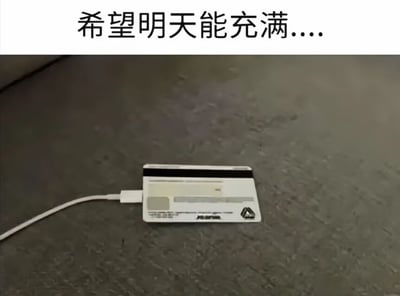 图片上方有一行文字“希望明天能充满....”，下方是一张信用卡平放在灰色纹理的表面上，信用卡上插着一根白色的充电线，仿佛在给信用卡“充电”。通过将信用卡与充电行为结合，幽默地表达了希望明天金钱充足的期待，带有生活调侃的意味。