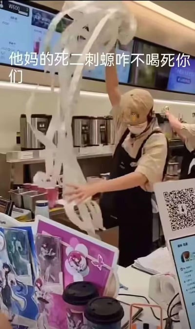 图片展示了咖啡店后厨场景，一名穿着黑色围裙的员工正高举手臂，手中大量白色奶油状物质被甩向空中，姿态夸张。画面上方叠加了醒目的白色文字：“他妈的死二刺螈咋不喝死你们”，语言极具攻击性。背景中可见咖啡机等设备，推测为奶茶店或咖啡店的工作场景。