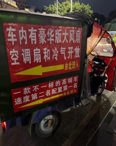 夜晚街头的一辆三轮车，车身两侧贴有醒目的红色大幅标语。上方标语写着“车内有豪华版大风扇 空调扇和冷气开放 由此进入”，配黄色箭头指向右侧；下方标语则称“一款不一样的高端车 速度第二名配置第一名 由此进入”，同样有黄色箭头。标语用词夸张，将普通三轮车宣传为“高端车”，以“豪华版”“配置第一名”等词汇制造反差幽默，展现了民间接地气的宣传风格。