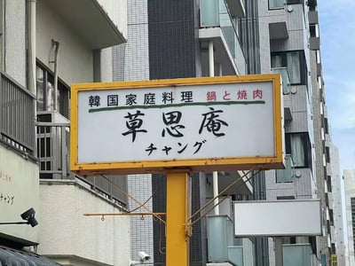 城市多层建筑背景下的韩国家庭料理店招牌，招牌为黄色边框，内有白色背景文字。上方文字为'韩国家庭料理 锅と烧肉'，中间是店名'草思庵'，下方有'チャング'字样。整体展现了城市中的异国料理餐厅标识。
