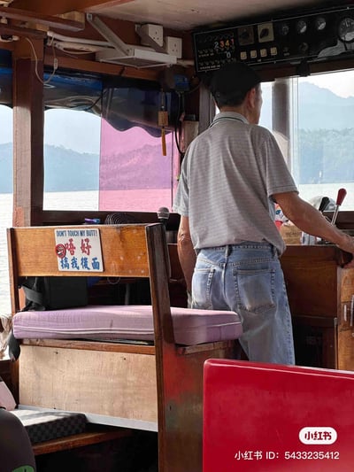 这是一张在船只驾驶舱内拍摄的真实场景图，一位老年男性船员背对着镜头，正专注操控船只，窗外是开阔的山水风光。前景的木质座椅靠背上，贴着一块充满趣味的双语标语牌：上方是英文“DON'T TOUCH MY BUTT!”，下方是粤语繁体“唔好搞我後面”，还搭配了一个禁止吸烟的图案。标语采用双关梗，字面意思是提醒不要触碰座椅后方区域，同时用谐音和直白的表述制造出滑稽的笑点，让普通的乘船场景变得趣味十足，充满接地气的生活幽默感。