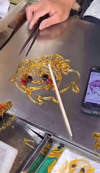 传统糖艺师傅正在金属台面上创作卡通角色糖画，金色糖浆勾勒出萌系人物轮廓，黑色糖豆做眼睛，红色糖粒点缀脸颊。右手持剪刀精细修整细节，旁边手机屏幕显示卡通原型图作为参考，台面上还散落着刀具等工具，展现传统手艺与现代卡通的创意结合。