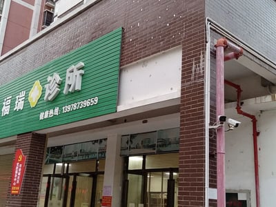 这是一张实地拍摄的社区临街诊所门店照片，建筑外墙采用红棕色砖面设计。门店上方的绿色招牌十分醒目，招牌上印有白色的“福瑞诊所”字样，搭配黄色的十字医疗标识，下方标注了健康热线13978739659。诊所入口为玻璃门窗结构，门窗上方贴有建筑类的装饰图片。在门店左侧墙面上，设有一个红色的指示牌，上面写着“华侨城社区党群服务中心 往前150米”，并带有党徽标识。建筑的右侧区域安装了两个监控摄像头，同时可见红色的消防管道和粉色的竖向管道，整体呈现出社区周边常见的线下医疗服务场所的真实样貌。
