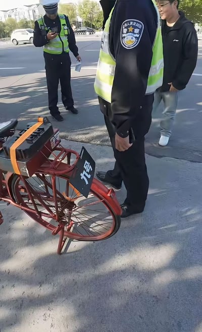 这是一张拍摄于城市道路旁的实拍照片，前景处是一辆经过私自改装的红色老式自行车，车架前端用橙色绑带固定了外置电瓶，前轮上方还挂着一块写有白色“九号”字样的黑色标识牌，模仿九号电动车的品牌标识。画面中有两名身着制式警服、外穿荧光绿反光背心的交通警察，其中一名站在改装自行车旁，另一名在稍远位置手持手机和纸质单据，正在处理该辆违规改装非机动车的相关事宜，一旁还站着一名穿黑色上衣、牛仔裤的年轻男子在围观，背景可见通行的机动车、道路绿化与远处的建筑物。