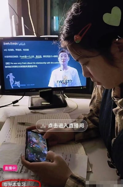 这张图呈现了充满反差感的备考场景：一名女生坐在桌前，桌面摆放着写有笔记的笔记本，身后的电脑屏幕正播放着B站的英语单词教学课程，看似在为专升本考试复习，但实际上女生正低头双手操作手机玩MOBA类手游，图中带有直播相关的界面标识，底部用红圈标注了“专升本复习”字样，讽刺了表面做学习准备、实则摸鱼玩乐的假性学习状态，很容易引发学生群体的共鸣。