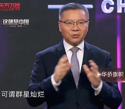 图片显示一位穿着深色西装、戴着眼镜的男性正在进行演讲，背景中可见东方卫视和《这就是中国》节目标识。人物双手做出演讲手势，表情严肃认真。画面底部有中文字幕“可谓群星灿烂”，右侧有“华侨旗帜”字样。