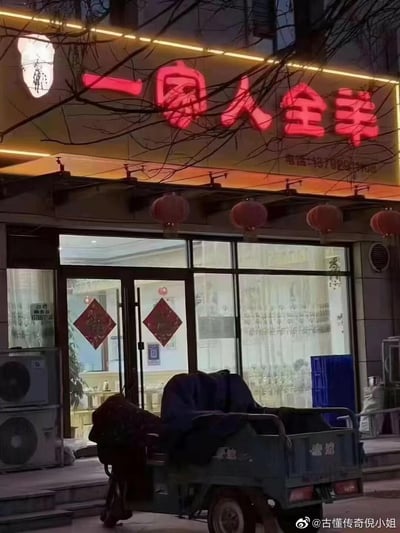 这是一张夜晚拍摄的街边餐馆实拍图，门店招牌为红色发光字体“一家人全羊”，下方标注了联系电话。门店的玻璃门和窗户上贴着倒贴的红色福字，门檐下悬挂着一排红色灯笼，门前停着一辆带有深色遮盖的电动三轮车。画面上方的树枝遮挡了部分招牌，整体呈现出温馨的日常街边餐饮门店夜景，没有双关、谐音梗等特殊内容。