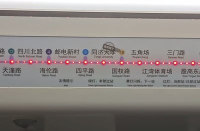 这是上海地铁10号线车厢内站点动态指示牌的实拍照片，指示牌分上下两行标注了线路途经的多个站点，上行标注了可换乘其他线路的站点及对应线路编号，包括四川北路（换乘12号线）、邮电新村（换乘4号线）、同济大学（换乘18号线）、五角场、三门路等，下行对应标注天潼路、海伦路、四平路、国权路、江湾体育场、殷高东路等站点；所有站点旁的指示灯均为红色，代表这些站点都已经被驶过；指示牌底部标注了友情提示，说明绿灯代表未驶过站、黄灯闪烁代表下一站、红灯代表当前站或已驶过站；最具趣味性的是在“同济大学”站点的上方，被人粘贴了一枚邮戳样式的贴纸，上面写有“卖不掉了”字样，形成调侃效果。