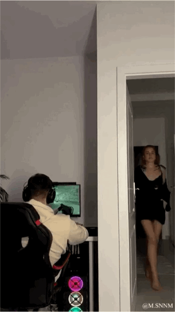 这是一段动态图（GIF），画面呈现居家场景：一名戴着头戴式耳机的男生正坐在电竞椅上，全神贯注地操作鼠标玩电脑上的射击类游戏，视线完全集中在电脑屏幕的游戏画面上；与此同时，一名身着黑色吊带短裙的女生从房间门口缓步走进来，男生似乎完全沉浸在游戏中，没有注意到女生的到来，整体展现出一种日常的居家生活反差感。