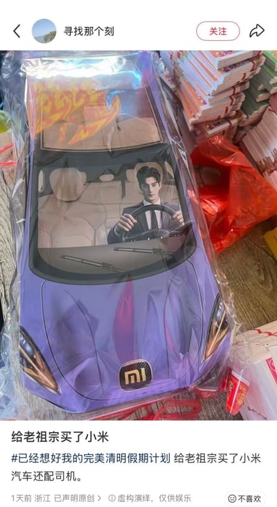 这是一张社交媒体帖子截图，主体是一款清明节祭祀用的纸扎汽车：纸扎汽车为紫色，车头印有小米品牌的“MI”标志，前挡风玻璃内的驾驶位印刷了一名穿西装打领带的男性，该男性双手握方向盘做出开车的姿态，纸扎汽车被透明塑料包装包裹，放置在一堆其他待售卖的纸扎纸品旁。帖子发布者昵称是“寻找那个刻”，配文表示这是给老祖宗买的小米汽车，还配备了司机，并标注内容为虚构演绎，仅供娱乐，发帖地点为浙江，发帖时间为1天前。