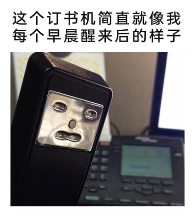 图片上方有一行黑色文字：“这个订书机简直就像我每个早晨醒来后的样子”。下方是一张实拍图，主体为一个黑色订书机，其顶部的金属压片部分因螺丝和凹槽的分布，恰好形成了类似人脸的图案——两个圆形螺丝孔像无神的眼睛，中间的椭圆形凹槽像鼻子，下方的横向凹槽则像微微下垂的嘴巴，整体呈现出一种疲惫、惺忪的表情。背景中隐约可见模糊的办公室电话和纸张，营造出日常办公环境的氛围。图片通过拟人化手法，将订书机的金属部件形态与人们早晨刚睡醒时的困倦状态联系起来，产生幽默共鸣效果。