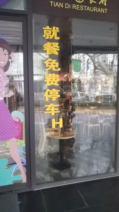 这是天地食府（TIAN DI RESTAURANT）门店玻璃上的实拍内容，玻璃上竖排张贴着黄色的“就餐免费停车H”提示文字，左侧还有手持购物袋的微笑卡通女性贴纸，玻璃同时倒映出店内的桌椅和店外的街道景象，是商家用来告知到店顾客可享受免费停车服务的实用提示，没有双关、谐音梗等特殊内容。