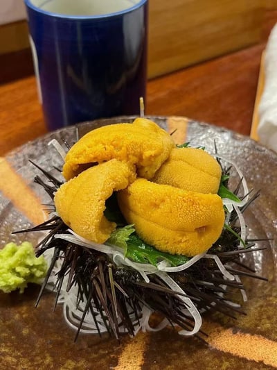 一张美食特写照片，展示了一盘新鲜的海胆刺身。金黄色的海胆黄被放置在保留着黑色尖刺的海胆壳中，周围点缀着绿色的紫苏叶和白色的细葱丝。盘子一侧配有一小团绿色的芥末。背景中可见一个深蓝色的杯子和木质桌面，整体呈现出日式料理的精致感。