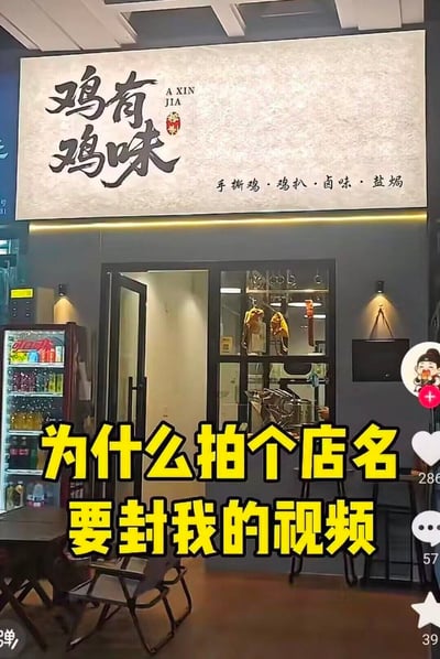 这是一张竖版的短视频平台截图，画面主体是一家餐饮门店的门面，门店上方的浅色招牌上用黑色书法字体写着店名「鸡有鸡味」，旁侧标注拼音「A XIN JIA」和红色印章样式标识，招牌右下角标注了门店经营品类「手撕鸡·鸡扒·卤味·盐焗」；门店左侧摆放着装满饮品的冷藏柜，门口有折叠桌椅，玻璃门内可以看到后厨悬挂的食材。画面下方叠加了醒目的黄底黑边大号文字「为什么拍个店名要封我的视频」，画面右侧显示了短视频平台的点赞、评论、收藏功能按钮及对应数据，还有创作者头像标识。该图的笑点在于店名「鸡有鸡味」本是表述鸡肉风味的正常内容，大概率被平台审核系统误判为违规内容，导致拍摄者发布的相关视频被封禁，从而引发拍摄者的吐槽。