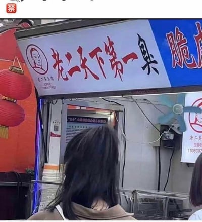 这是一张街头小吃摊位的实拍静态图，画面上方是蓝白配色的摊位招牌：招牌左侧印有一个卡通人物头像，中间用醒目的红色大字写着“老二天下第一臭”，下方配有小字“老二臭豆腐”和联系电话“15063071405”，右侧蓝色背景区域有白色的“脆皮”字样。摊位前能看到两位顾客的背影，其中一位长发女生位于画面下方，摊位内部可以看到张贴的相关经营证照，还挂着一个小风扇，摊位左侧悬挂着红色的灯笼，整体呈现出热闹的街头美食场景。招牌用夸张的“天下第一臭”来突出臭豆腐的特色，没有双关、谐音梗或暗示内容。