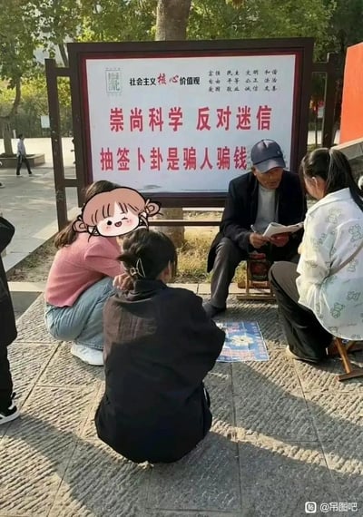 画面呈现了极具讽刺性的一幕：在一块明确写着“崇尚科学 反对迷信 抽签卜卦是骗人骗钱”的社会主义核心价值观宣传牌前，一位算命先生正坐在小凳上，拿着纸笔给几位围坐的年轻人算命，宣传牌的内容和眼前的行为形成强烈反差，充满讽刺感。