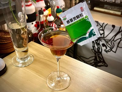这是一张在酒吧吧台拍摄的创意整活图，将白云山感冒灵颗粒的包装袋插在一杯盛有琥珀色液体的调酒杯边缘，营造出“喝感冒灵调酒”的搞怪效果。背景中可以看到带有圣诞风格装饰的酒瓶、插着水培植物的玻璃容器，整体场景轻松有趣，是将日常药品与酒吧调酒结合的创意恶搞，没有双关或谐音梗，纯靠实物摆放的创意制造笑点。