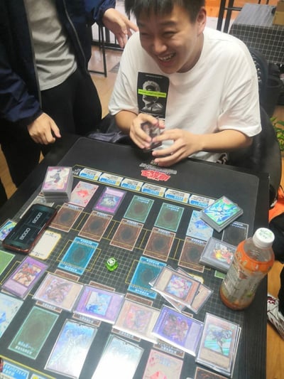 这是一张原始实拍照片，记录了线下桌游聚会的轻松场景。画面中心是一位穿着白色印花T恤的男性玩家，他正开怀大笑，情绪十分兴奋，双手握着卡牌，似乎在整理或准备进行卡牌操作。桌上铺着《游戏王》的官方对战垫，上面摆放着多组不同的游戏王卡牌，还有一部手机、一瓶橙色瓶装饮料，旁边另一位站着的人正关注着对局，整体氛围愉快，展现了卡牌爱好者享受游戏的瞬间。