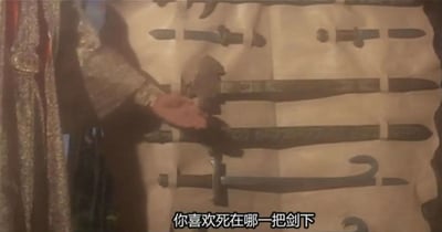 这是一张经典香港武侠电影《新龙门客栈》的影视截图，画面中展示了铺展在桌面上的数把冷兵器，涵盖短剑、长刀等不同形制，一只手正搭在其中一把兵器的柄部，画面下方配有字幕“你喜欢死在哪一把剑下”，瞬间营造出剑拔弩张、充满压迫感的对峙氛围，是影片中极具江湖凶险感的挑衅名场面，将武侠世界的紧张冲突感拉满。