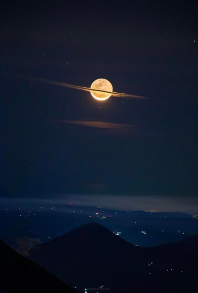 这是一张夜晚自然景观照片，画面中央是一轮明亮的满月，月光皎洁，周围环绕着稀疏的星星。一道纤细的云层横向穿过月亮，增添了画面的层次感。下方是深色的山脉轮廓，山脚下可以看到远处城市的灯光点缀，形成了自然景观与人类文明的和谐对比。整个画面呈现出宁静而壮丽的夜空景象。
