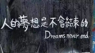 这是一张静态的涂鸦类标语图，画面背景是带有自然纹理和水渍痕迹的深色木板，整体色调暗沉，带有粗糙的质感。木板上用白色颜料手绘出两行文字，上方是简体中文“人的梦想是不会结束的”，下方是对应的英文翻译“Dreams never end”，白色字体笔触粗犷，与深色背景形成鲜明对比，清晰传递出鼓励人们坚持梦想、永不言弃的励志情绪。
