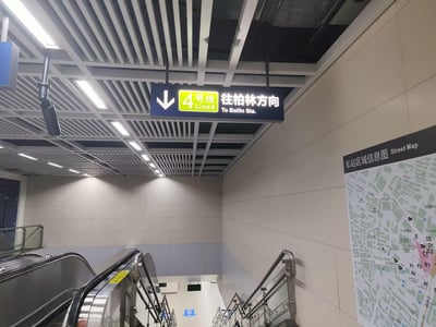 这是一张武汉地铁车站内的实拍照片，画面中天花板悬挂着一块指引标识，标注着“4号线 Line4 往柏林方向 To Bailin Sta.”，下方箭头指向通往乘车区域的自动扶梯与楼梯；右侧墙面张贴着车站区域信息图，展示车站周边的道路与出入口分布。整体环境整洁明亮，为乘客提供明确的乘车方向引导，是典型的城市轨道交通站内场景。