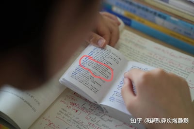 这是一张记录学习日常的实拍图，画面中一只手握着一本小型英语词汇手册，手册上用红圈重点标注出deceit、deceive、deceitful等英语单词及对应的中文释义、搭配用法；手册下方是一本写满红色手写数学公式的专业书籍，背景还能看到堆叠的其他书本，整体展现出同时进行英语词汇积累与数学学习的专注学习状态。