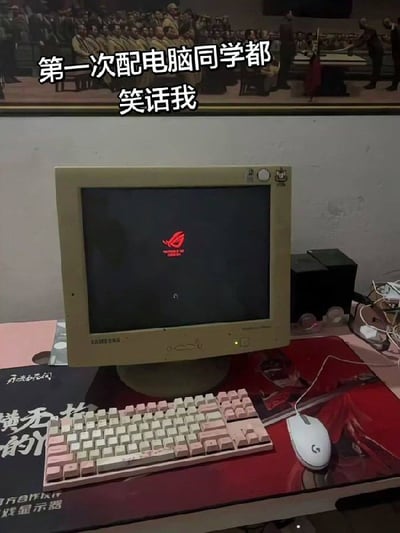 整张图拍摄了桌面的一套电脑设备，使用已经明显泛黄老旧的复古CRT显像管显示器，但屏幕上却显示着华硕高端电竞品牌ROG（玩家国度）的开机标识，显示器前方搭配了粉白配色的机械键盘、罗技高端电竞鼠标，桌面铺有电竞相关桌垫。图片上方叠加了白色文字“第一次配电脑同学都笑话我”，通过外观老旧的显示器和高端内部配置的强烈反差制造笑点，调侃他人以貌取机、不懂实际配置高低的情况。
