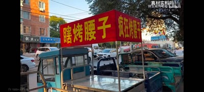 这是一张充满市井生活气息的街头实拍图，画面核心是一辆停放在城市路边的三轮小吃摊，摊位上方的红色招牌用醒目的黄色字体写着“瞎烤腰子”，招牌侧面则标注着“疯狂烤腰子”，这种自黑式的搞笑招牌形成了有趣的反差感，十分吸睛。背景中可以看到多辆停放的私家车、居民住宅楼，还有街边的“靓丝造型”理发店与“蓝色经典·天之蓝”烟酒商店，整体场景还原了日常的城市街头风貌，充满生活烟火气，招牌的趣味命名很容易引发路人与网友的调侃。